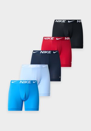 Skjortlös man som står barfota, klädd i blå Nike boxerkalsonger, poserad med ena handen bakom ryggen mot en enfärgad ljus bakgrund.