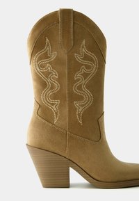 Zamszowy, zachodni but w kolorze koniakowym, z zaostrzonym noskiem, ukośnym obcasem i ozdobnym przeszyciem w kształcie wirującego wzoru na cholewce.