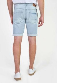 Man in lichtblauwe opgerolde denimshorts, witte sneakers en een wit shirt, gezien van achteren tegen een effen achtergrond.