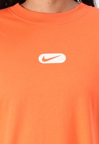 Oranža kokvilnas T-kreklā ar baltu Nike logo centrā. Audums ir gluds, ar klasisku apaļo kakla izgriezumu un īsām piedurknēm.
