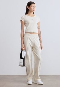 Κρεμ ριμπ τοπ, φαρδιά κρεμ παντελόνια με κορδόνι, λευκά αθλητικά παπούτσια, ασημένια τσάντα με μαύρη λαβή, μινιμαλιστικό στυλ, ομαλές υφές.