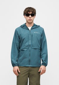 Άνδρας που φοράει τιρκουάζ φούτερ με κουκούλα Columbia, μαύρα γυαλιά ηλίου, χακί παντελόνι cargo, στέκεται μπροστά σε απλό λευκό φόντο.