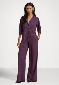 Lilla mønstret jumpsuit med krage, lange ermer, elastisk midje og vide ben. Glatt stoff med geometrisk trykk.