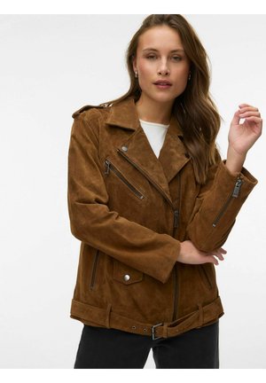 Femme portant une veste de motard en suède marron avec des poches zippées et une ceinture, posant les bras croisés devant un fond blanc uni.