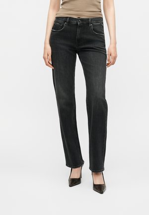 Frau trägt schwarze Jeans mit geradem Schnitt, beiges Top und schwarze spitze High Heels, steht vor einem schlichten hellen Hintergrund.