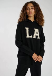 Svart hoodie med lös passform, med en stor krämfärgad "LA"-logotyp på framsidan. Tillverkad av mjukt tyg, med ribbade muddar och nederkant.
