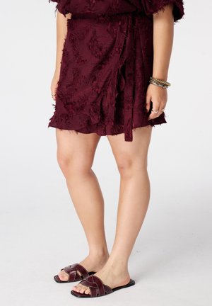 Vrouw draagt een gestructureerde bordeaux wrapjurk met bijpassende platte slip-on sandalen en armbanden om haar rechterpols tegen een witte achtergrond.