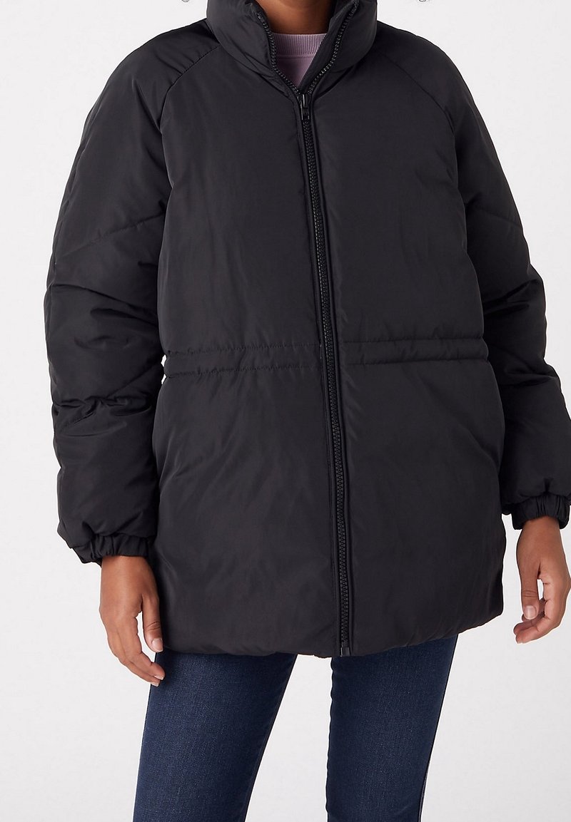 Personne portant une veste d’hiver noire matelassée avec une fermeture éclair frontale, des poignets élastiques et un col montant, associée à un jean bleu foncé.