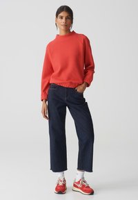 Rode textuurtrui met een hoge hals, gecombineerd met donkere denim wijde pijpen spijkerbroek. De outfit bevat witte enkel sokken en rode sneakers.