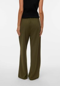 Persoon draagt olijfgroene wijde broekspijpen en een zwart mouwloos topje, staand met ontspannen handen langs het lichaam tegen een witte achtergrond.