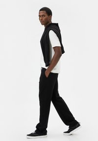 Pantalones de chándal negros, camiseta blanca, sudadera negra caída sobre los hombros, zapatillas negras. El modelo está de perfil sobre un fondo blanco liso.