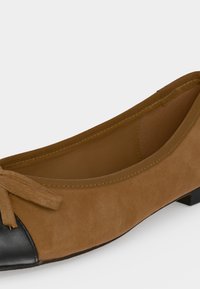 Ballerine en daim marron avec un bout arrondi noir et un petit nœud sur le dessus.