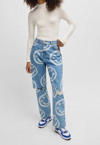 Jasnoniebieskie dżinsy z denimu z powtarzającym się białym wzorem uśmiechniętej buźki, przetarciami i luźnym krojem. Do zestawu biała prążkowana bluzka i trampki.