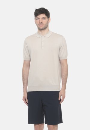 Uomo in piedi, indossa una polo beige a maniche corte e pantaloncini neri fino al ginocchio su uno sfondo bianco uniforme.