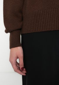 Καφέ πλεκτό πουλόβερ με ριμπ μανσέτες και τελείωμα, εμφανιζόμενο με το χέρι να ακουμπά σε μαύρο ύφασμα. Έμφαση στην υφή και την αντίθεση χρωμάτων.