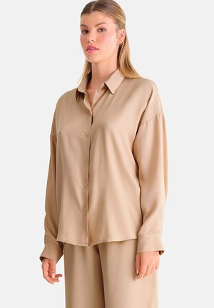 Vrouw draagt een loszittend beige overhemd met knopen en bijpassende broek, staand tegen een effen lichte achtergrond.