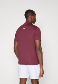 Burgundska polo majica s ovratnikom, kratkimi rokavi in belim logotipom na hrbtni strani. Gladko bl material in prilagojen kroj, v kombinaciji z belimi kratkimi hlačami.