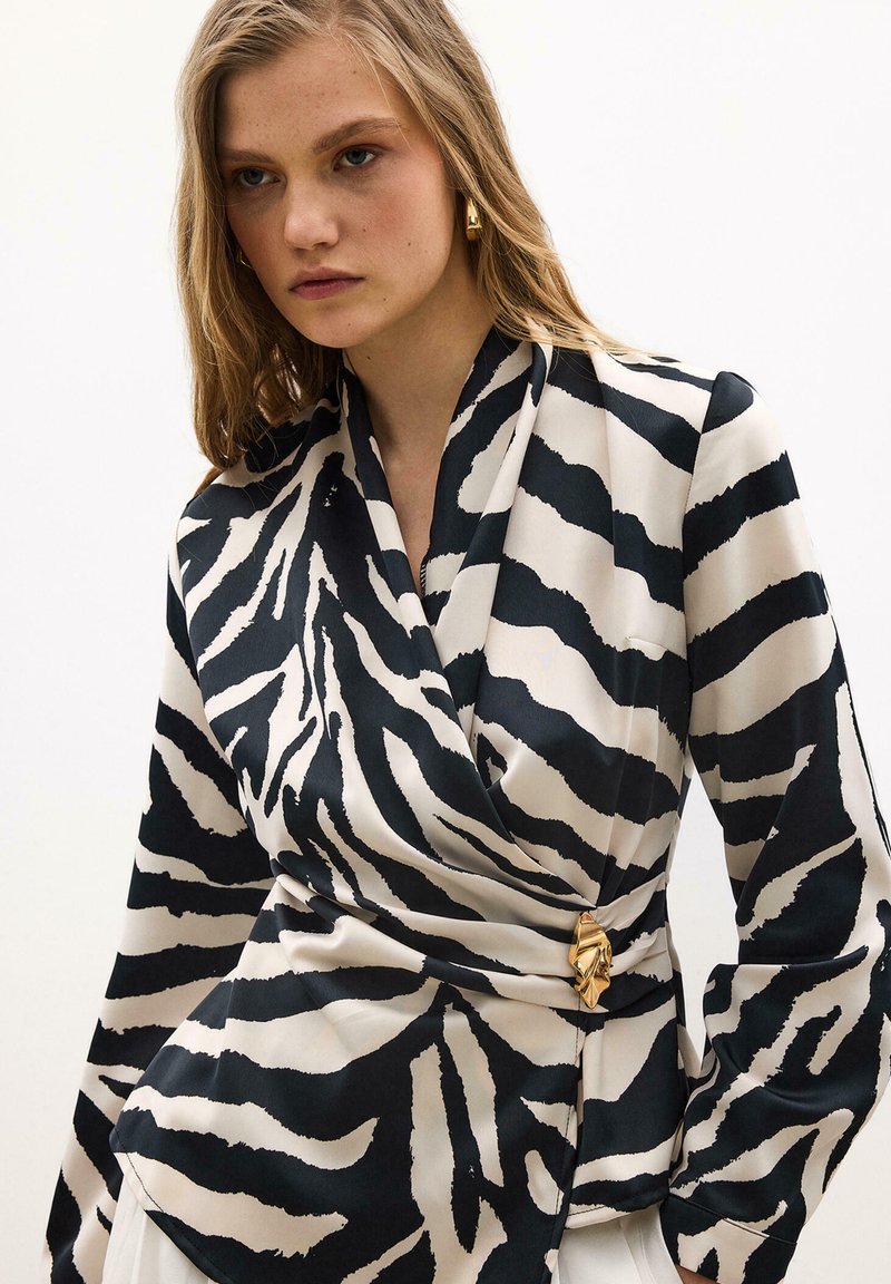 Jeune femme portant un chemisier cache-cœur à imprimé zèbre noir et blanc avec une boucle dorée et des boucles d'oreilles créoles, regardant légèrement vers le bas.
