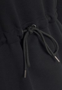 Svart tyg med en texturerad, elastisk midja. Har en dragsnodd med metalldetaljer och en knuten detalj som förbättrar greppet.