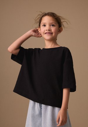 Ung pige med løst hår iført en sort oversize skjorte og lyseblå jeans, som kigger lidt opad med en hånd, der rører ved hendes øre.