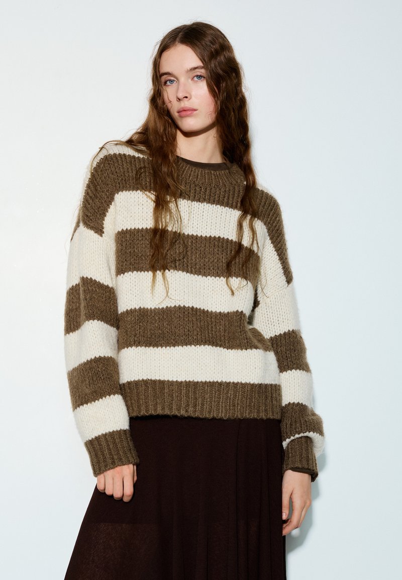 Pull rayé en maille beige et brun foncé, avec un ourlet et des poignets côtelés, associé à une jupe foncée. Manches longues et coupe décontractée.