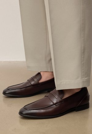 Braune Ledermokassins getragen mit hellbeigen, maßgeschneiderten Hosen auf einem glatten Boden.