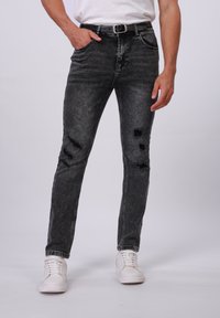 Jeans de mezclilla negra con un corte ajustado, que presentan parches desgastados, un ligero desvanecimiento, cinco bolsillos, y combinados con zapatillas blancas.