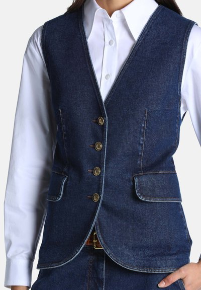Femme portant un gilet en denim bleu foncé avec quatre boutons et poches à rabat sur une chemise blanche boutonnée.