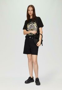 Schwarzes Grafik-T-Shirt mit kreisförmigem Design, schwarze Baumwollshorts und schwarze Sneaker. Das Model hält eine kleine schwarze Tasche mit einer Kettenriemen.