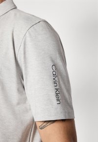 Lysegrå poloskjorte med korte ermer og krage. Har brodert "Calvin Klein" tekst på venstre erme. Mykt, teksturert stoff.