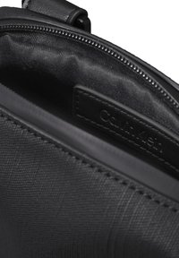 Interior negro texturizado de un bolso con cierre de cremallera y una etiqueta con "Calvin Klein" en relieve. Superficie de material sintético suave.