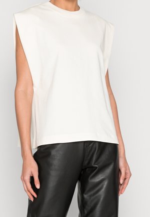 White sleeveless padded shoulder top paired with loose black leather pants on a standing person against a plain background.