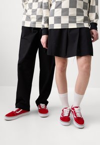 Baskets rouges avec des lacets blancs, portées par deux personnes. L'une porte une jupe plissée noire, l'autre un pantalon noir, les deux portent des hauts à motifs.