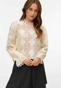 Koronkowa bluzka z falbaniastymi kołnierzykiem i mankietami w kolorze kremowym, zdobiona geometrycznymi wzorami i długimi rękawami, w zestawieniu z ciemną spódnicą.