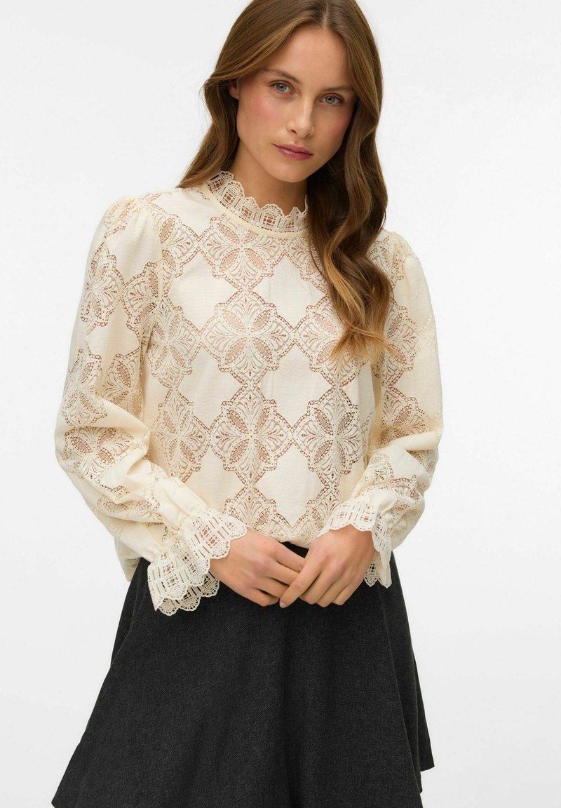 Koronkowa bluzka z falbaniastymi kołnierzykiem i mankietami w kolorze kremowym, zdobiona geometrycznymi wzorami i długimi rękawami, w zestawieniu z ciemną spódnicą.