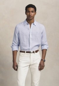 Νεαρός άνδρας που φοράει ένα ανοιχτό μπλε πουκάμισο με γυρισμένα μανίκια και λευκά παντελόνια, στέκεται μπροστά σε ένα απλό μπεζ φόντο.