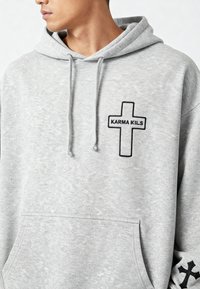 Felpa grigia con cappuccio realizzata in tessuto morbido, caratterizzata da un logo centrale a croce nera con la scritta "KARMA KILS" e ulteriori decorazioni a croce nera sulle maniche.