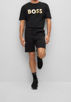 Man wearing black t-shirt with gold "BOSS" text, black shorts, black socks, and black sneakers, standing with one leg raised against gray wall.