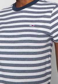 Man som bär en vit och marinblå randig t-shirt med marinblå krage och en liten rektangulär logotyp på bröstet.