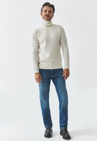 Pulover alb cu guler înalt, având un design ajustat, asortat cu blugi albaștri și pantofi negri cu șireturi. Fără modele sau decorațiuni distincte.