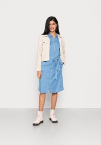 Ljusblå denimklänning med knytmidja, knapprad fram och två bröstfickor, lagd under en krämfärgad kort jacka och parat med rosa stövlar.