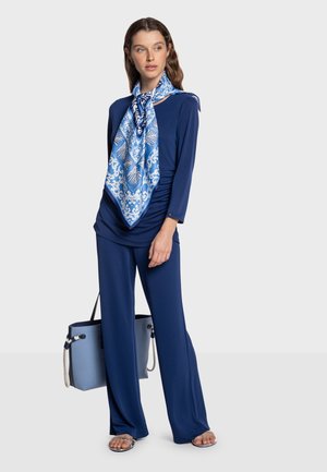 Haut bleu marine avec un design drapé, associé à un pantalon large assorti. Un foulard bleu à motifs est noué autour du cou. Complété par un sac à main bleu.