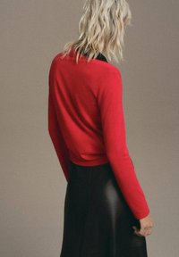 Haut rouge à manches longues associé à une jupe en cuir noire, mettant en valeur la texture lisse du vêtement et son design ajusté vu de l'arrière.