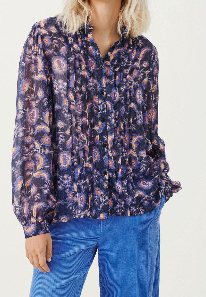 Femme portant un chemisier boutonné sombre à motif floral avec des plis, associé à un pantalon en velours côtelé bleu vif, debout avec une main dans la poche.