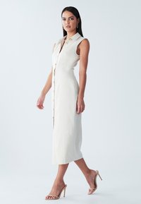 Roomkleurige, mouwloze jurk met knopen en kraag, met een rechte snit, halflang en een natuurlijke stoftextuur. Eenvoudige houten knoopdetails.