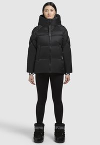 Black puffer jacket with a high collar, zip front, and boxy shape. Features quilted padding and two front pockets. Worn with black leggings and boots.