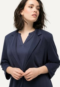 Marinblå blazer med mjuk textur, snedskurna slag och trekvartslånga ärmar, kombinerad med en marinblå V-ringad topp. Modellen justerar knappen på blazern.