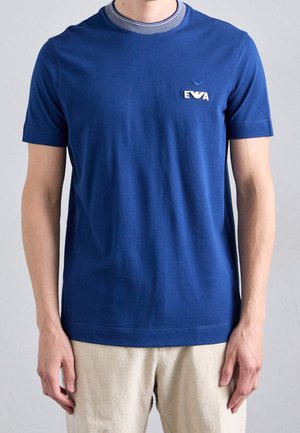 Man draagt een blauw T-shirt met korte mouwen, gestreepte kraag en wit logo op de borst, gecombineerd met lichtbeige broek.