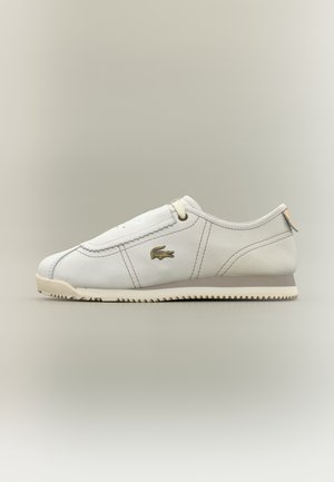 Zapatilla blanca de corte bajo con suela beige, detalles cosidos, logo del cocodrilo de Lacoste en el lateral y cordones mínimos sobre un fondo liso.