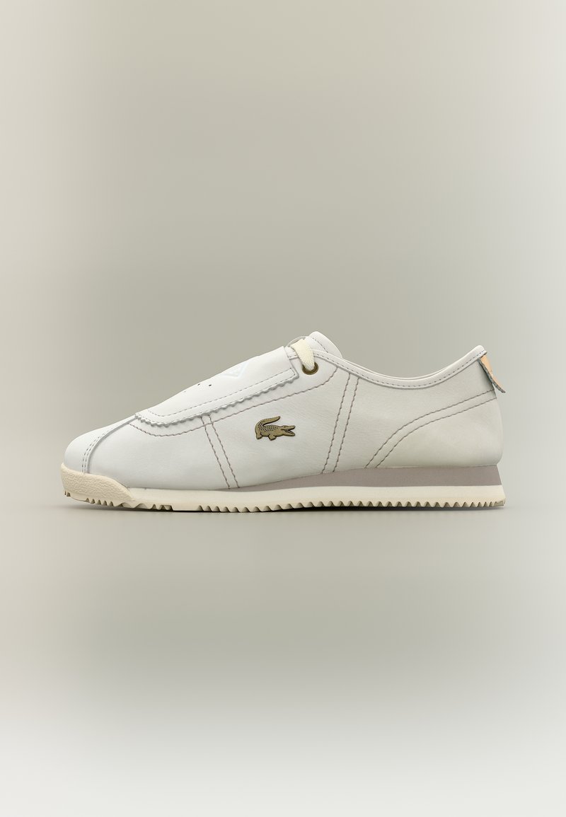 Λευκό sneaker χαμηλού ύψους με μπεζ σόλα, ραμμένες λεπτομέρειες, λογότυπο κροκοδείλου Lacoste στο πλάι και ελάχιστα κορδόνια σε απλό φόντο.